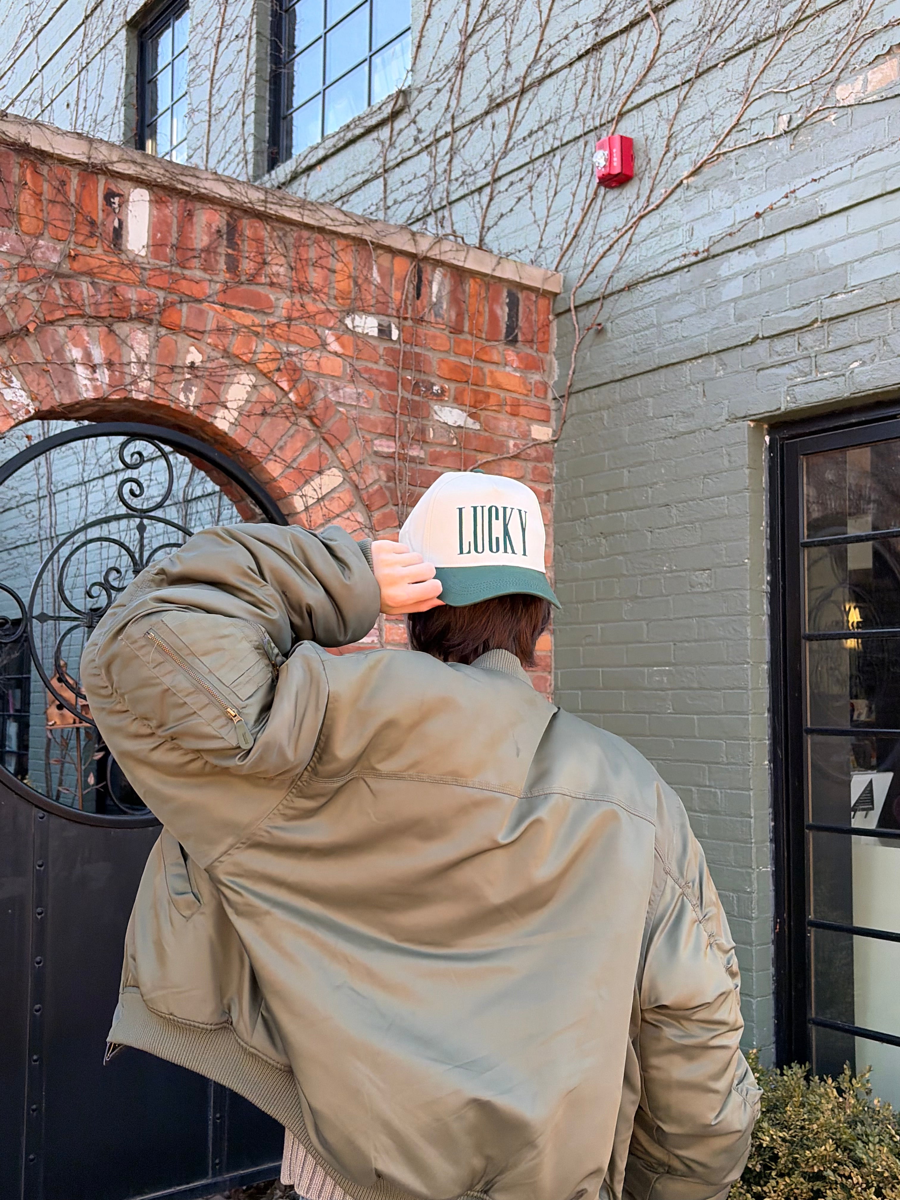 Person wearing a beige jacket and 'Lucky' cap in front of a brick and gray building.
