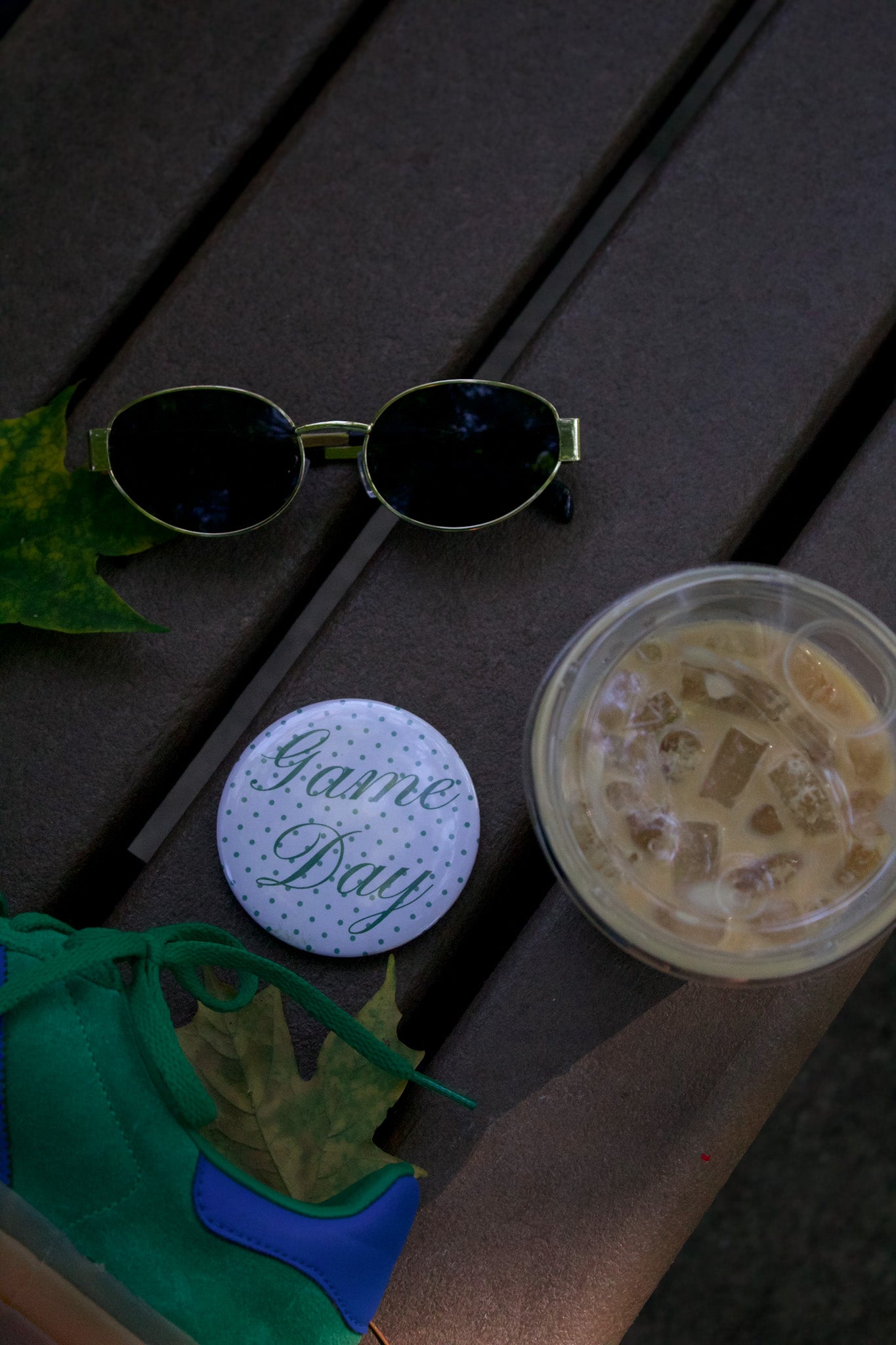Sunglasses, 'Game Day' coaster, and cup on a wooden surface with leaves.