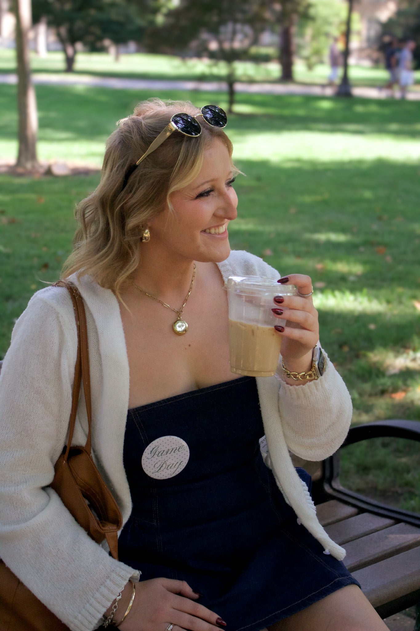 Woman sitting on a park bench holding a drink, wearing sunglasses on her head and the "Game Day" Button.